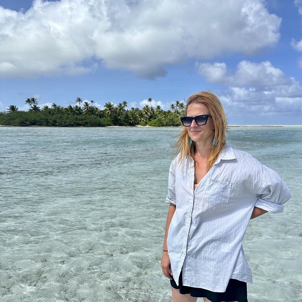Carla Steele, wearing a light blue blouse, long orange hair and sunglasses on the beach with an island in the background.
