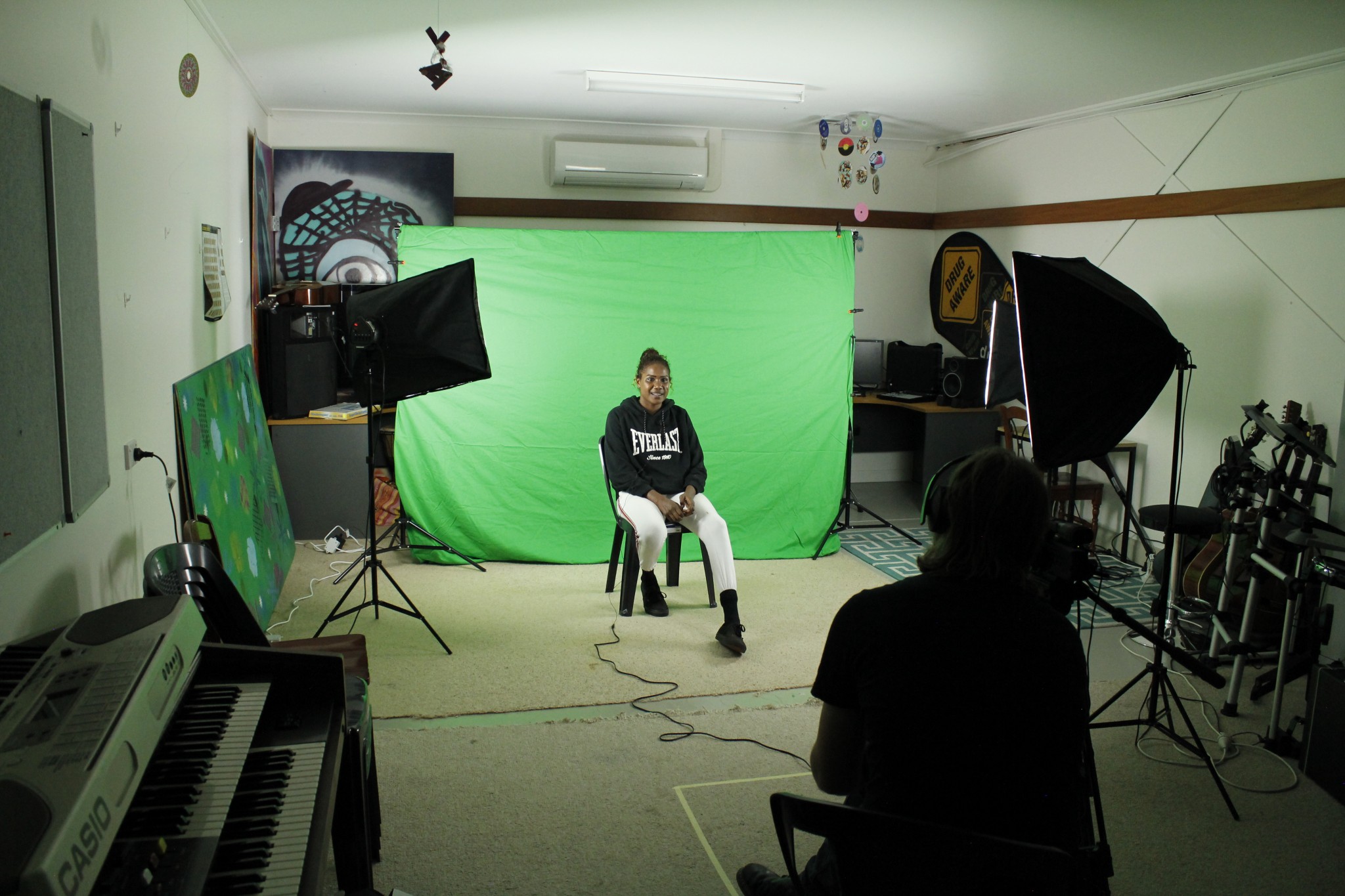 Person sits on chair in front of green screen with lighting boxes in front of them and a person filming.