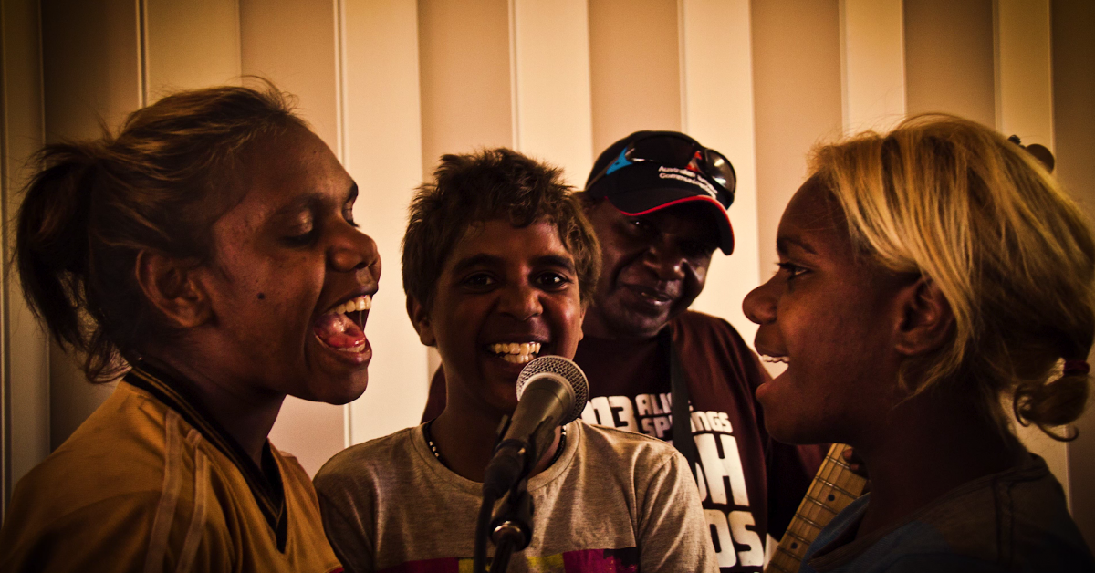 Three people sing into a microphone, 1 person watches.