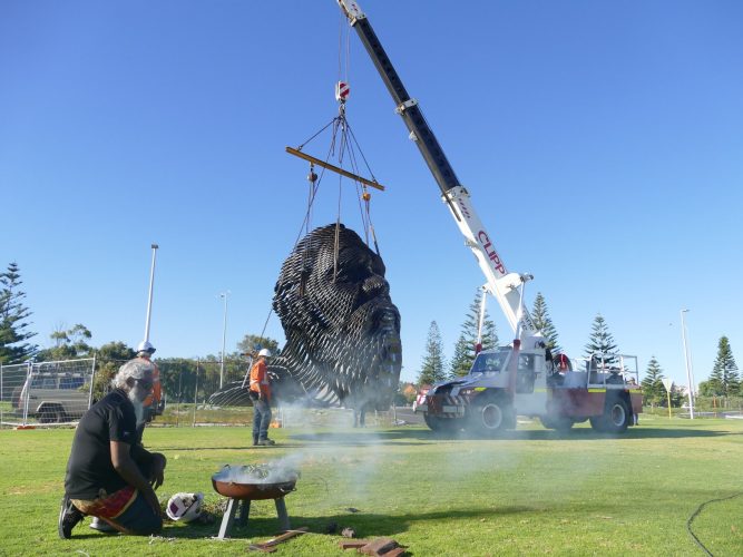 A smoking ceremony takes place in front of a giant sculpture of a head being lifted into the air by a truck.