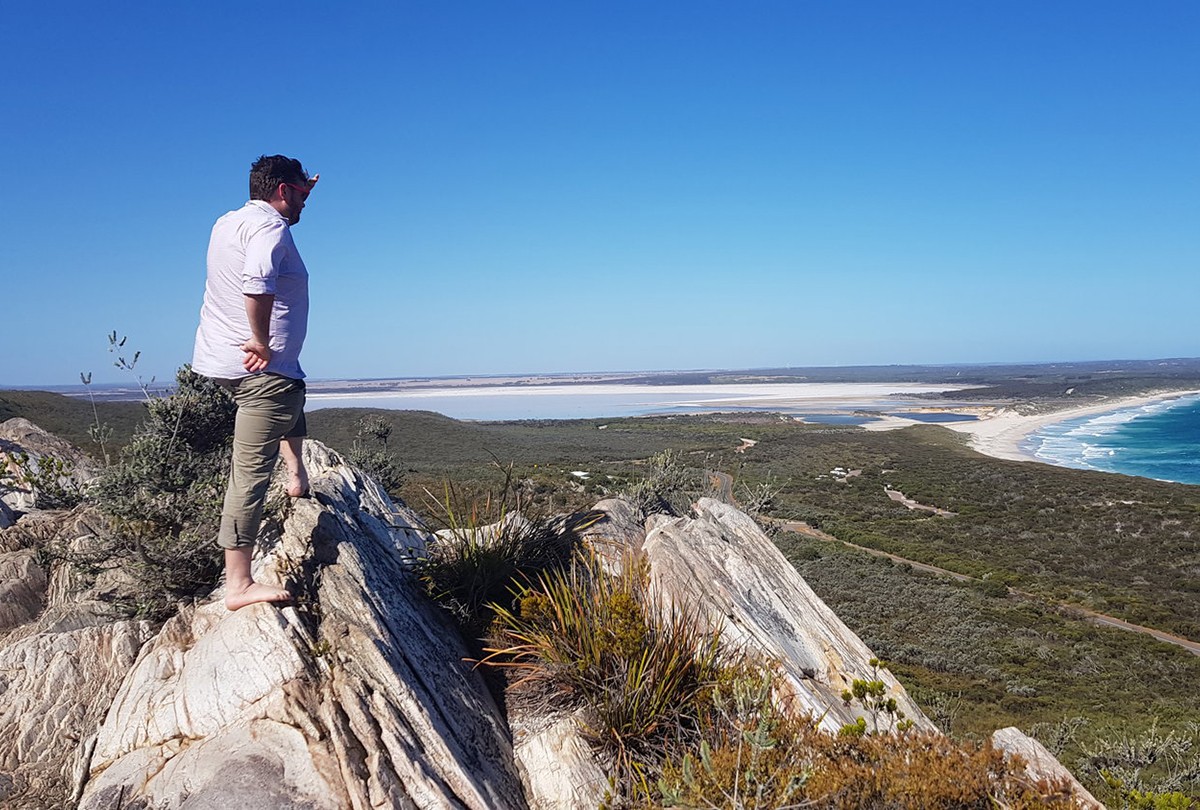 Barefoot man stands on rocky ledge overlooking an inlet meeting the ocean.