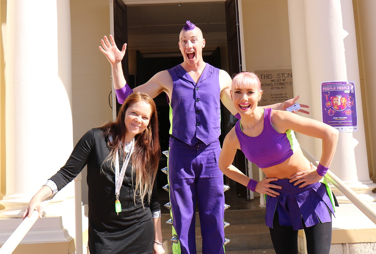 Three people standing for a photo outside a building, two wear purple, the other is dressed in black.