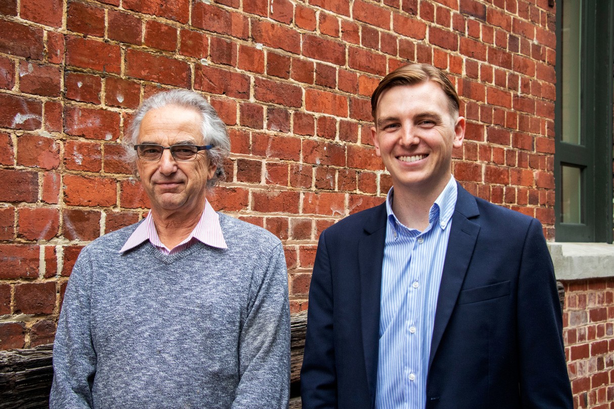 Two people stand in front of a brick wall; one in a grey sweater and collared shirt, the other in a dark suit and blue shirt.