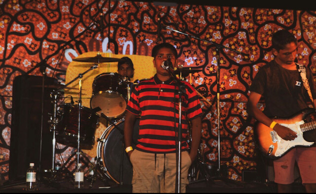 Youth band performs on stage, one boy has the microphone, another plays drums and the third plays the electric guitar.