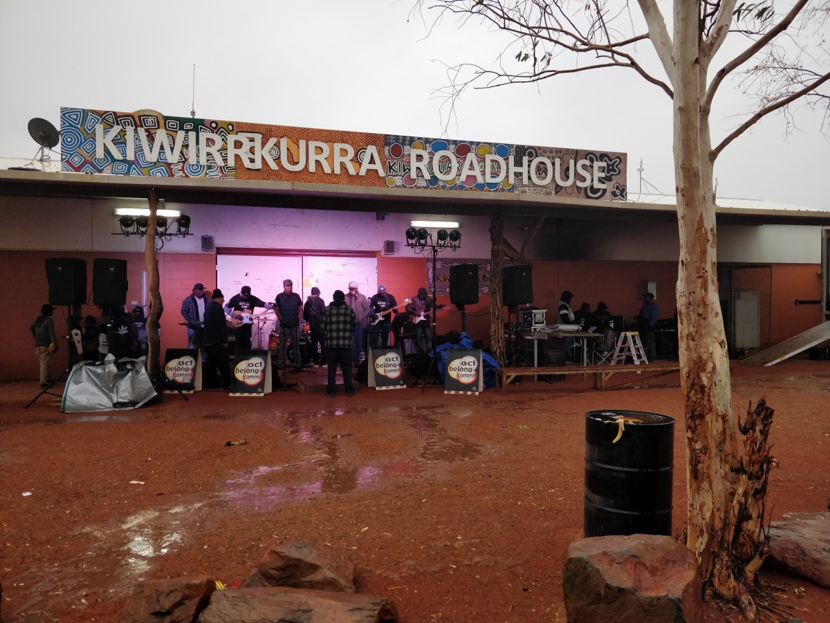 Outdoor stage at “Kiwirrkurra Roadhouse” with a band performing; wet ground, rocks, tree in foreground, and colorful sign above the stage.