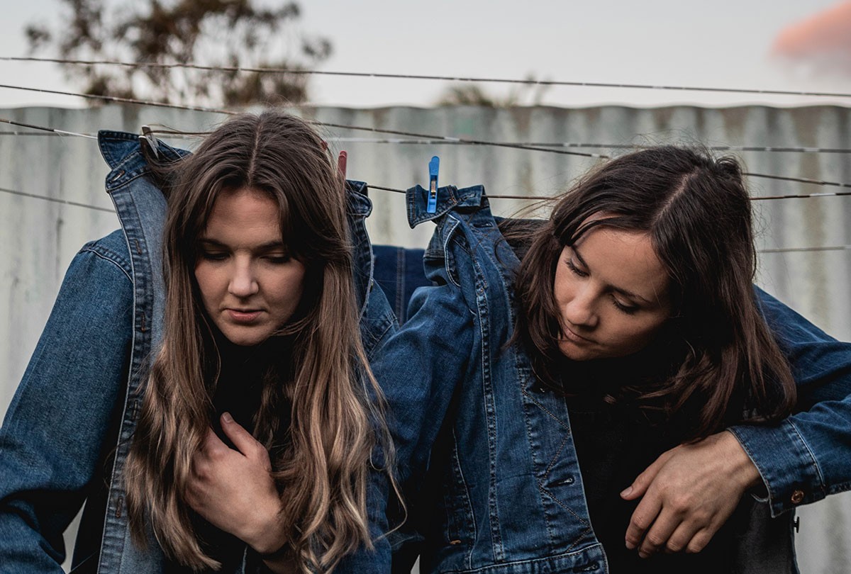 Two individuals with long hair in denim jackets hang by their collars on a clothesline with clothespins.