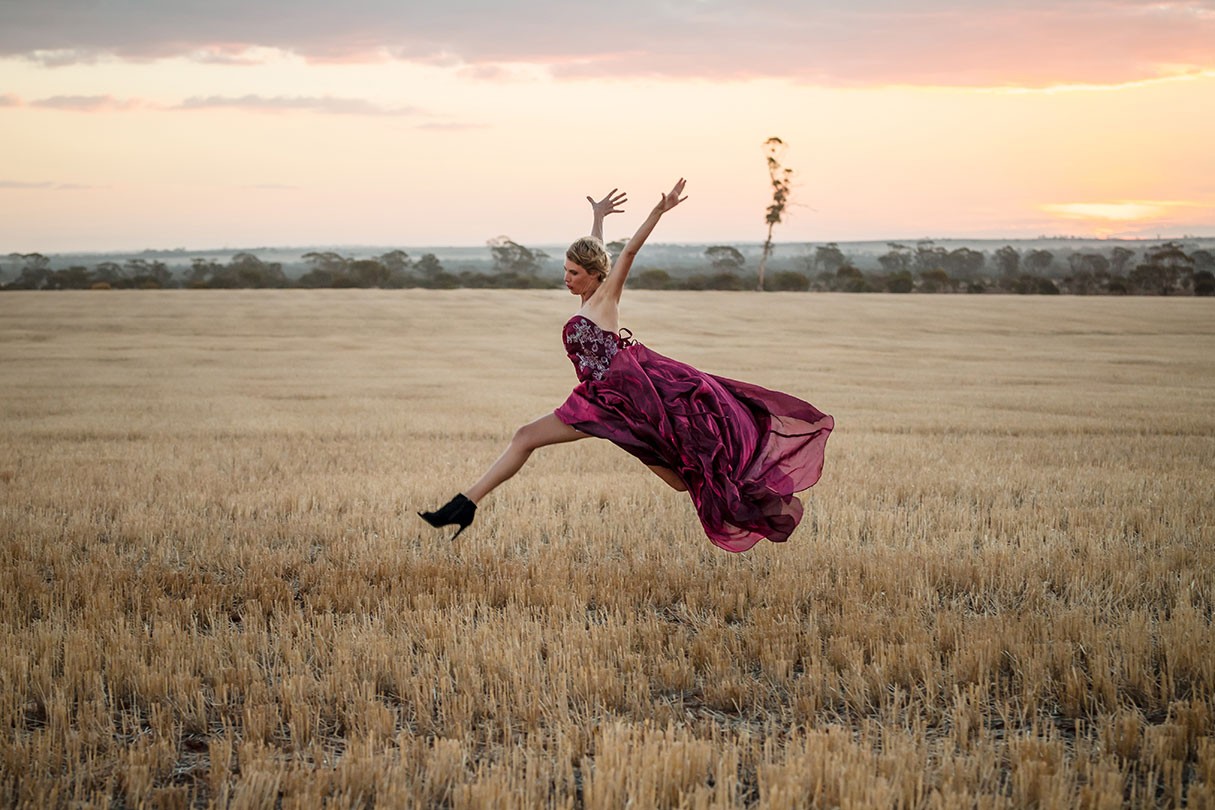 Person leaps through harvested field wearing a flowy pink strapless dress and heeled ankle boots.