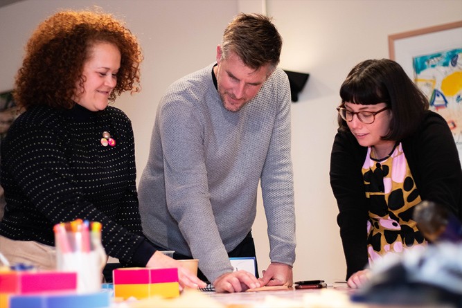 Three people look intently at a piece of paper set out between them. Two of them point to different parts of the paper as they converse.