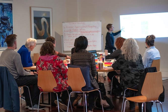 A woman stands in front of a small crowd of six or so people, pointing to something on a projector screen. A whiteboard stands to the left of the presenter, filled with writing.