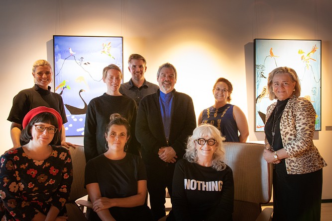 A group of people stand smiling in front of a brightly lit gallery wall, adorned with two paintings. Each painting is blue in hue, and depicts native birds.