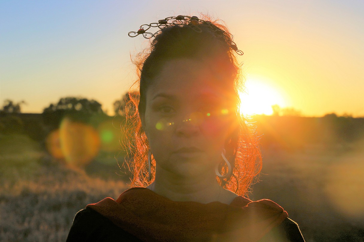 A picture of artist, Yola Baker, with the sun setting behind her. She is in partial silhouette, obscured by glare.