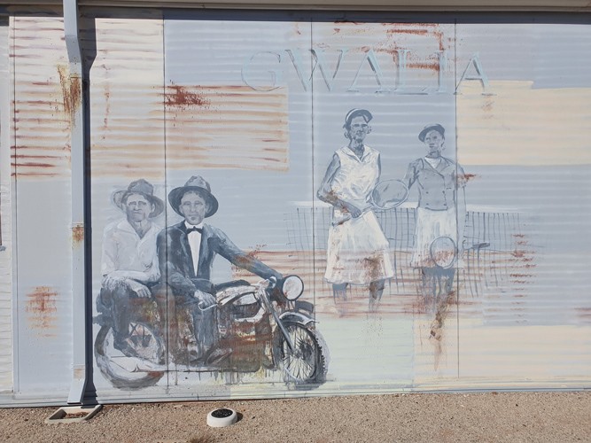 A mural on a tin wall, depicting a pair of women (dressed in attire which looks dated - perhaps late 60's) preparing for a tennis match and a man on a motorbike (dressed quite formally - including suit, bow tie, and bowler hat) - another man sits on the back of his bike catching a ride. The word "Gwalia" can be made out faintly in the background of the mural.