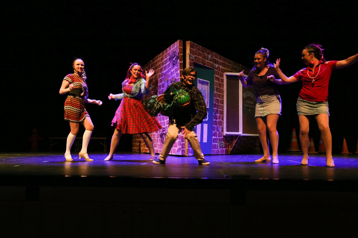 Stage performance with five people around a central figure holding a large green plant-like prop, set against a brick building backdrop.