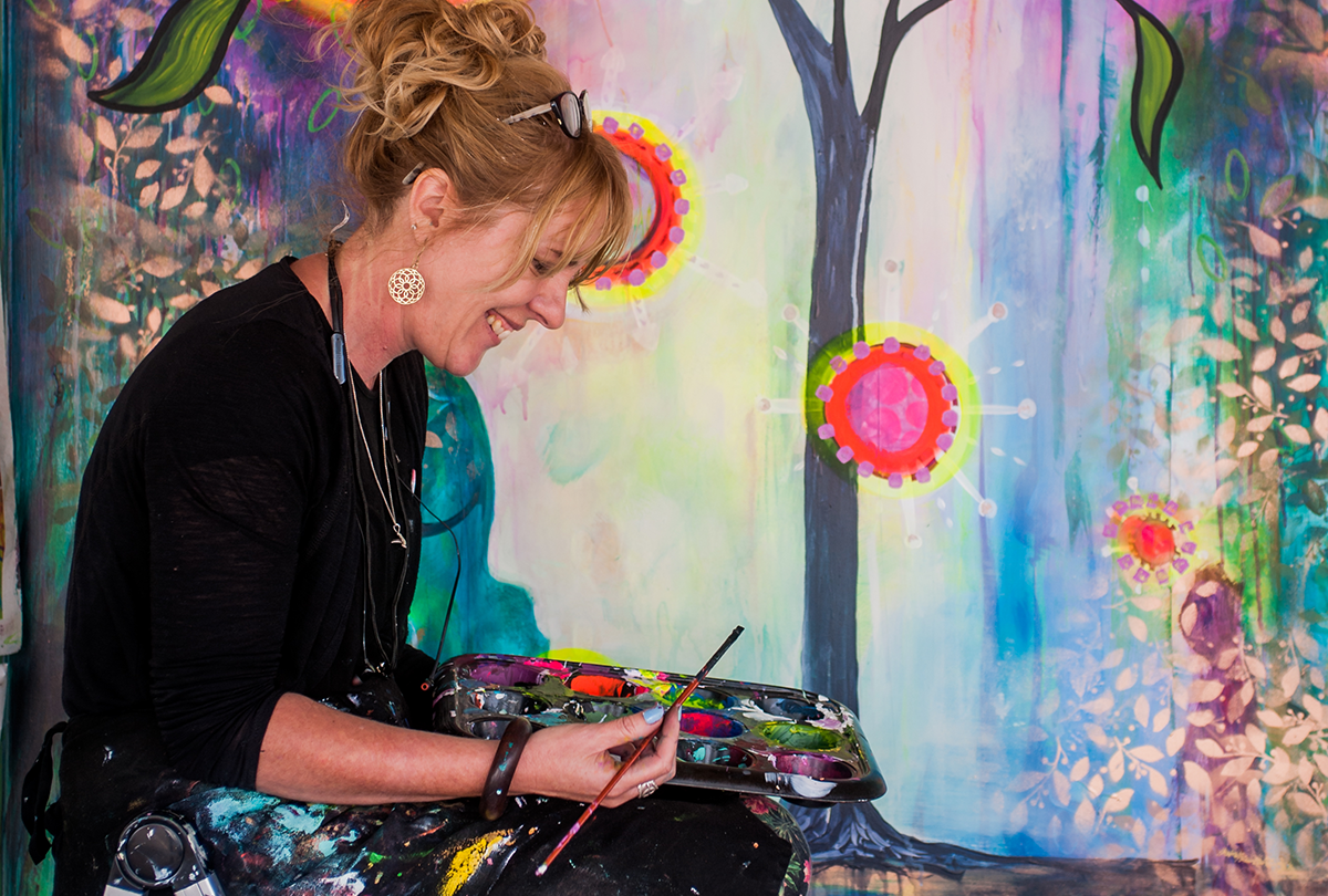 Artist holds paintbrush and pallet of paints, in front of a large artwork.