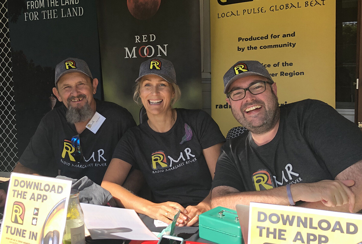 Three people sit at a table with RMR promo items, wearing branded gear; banners for Voyager Estate, Red Moon, and RMR behind them.