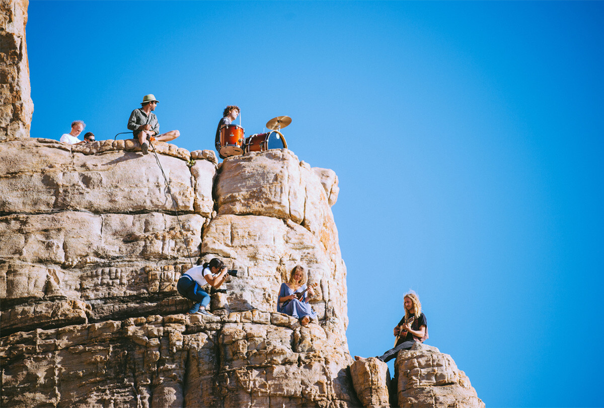 Seven people on a rocky cliff under a clear blue sky—one plays drums, one takes photos, others sit or stand at different levels.