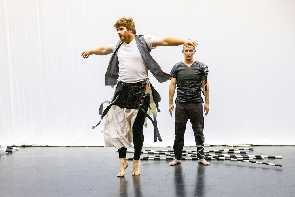 Two people in grunge costumes dancing, one with arms outstretched, the other behind in front of a pile of black and white poles.