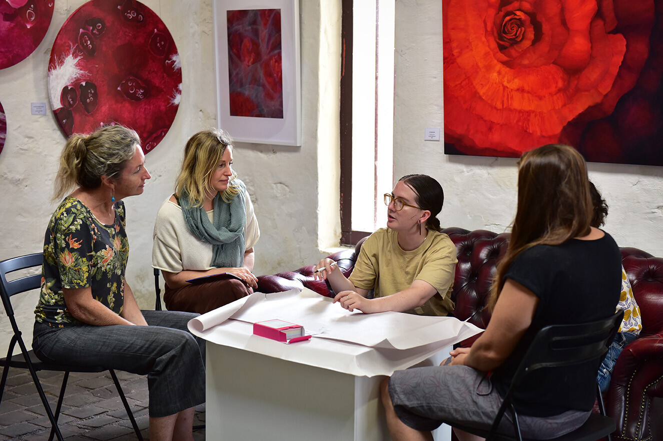 Five people in a room with red artworks and a red couch, they are seated around a small table with paper on it.