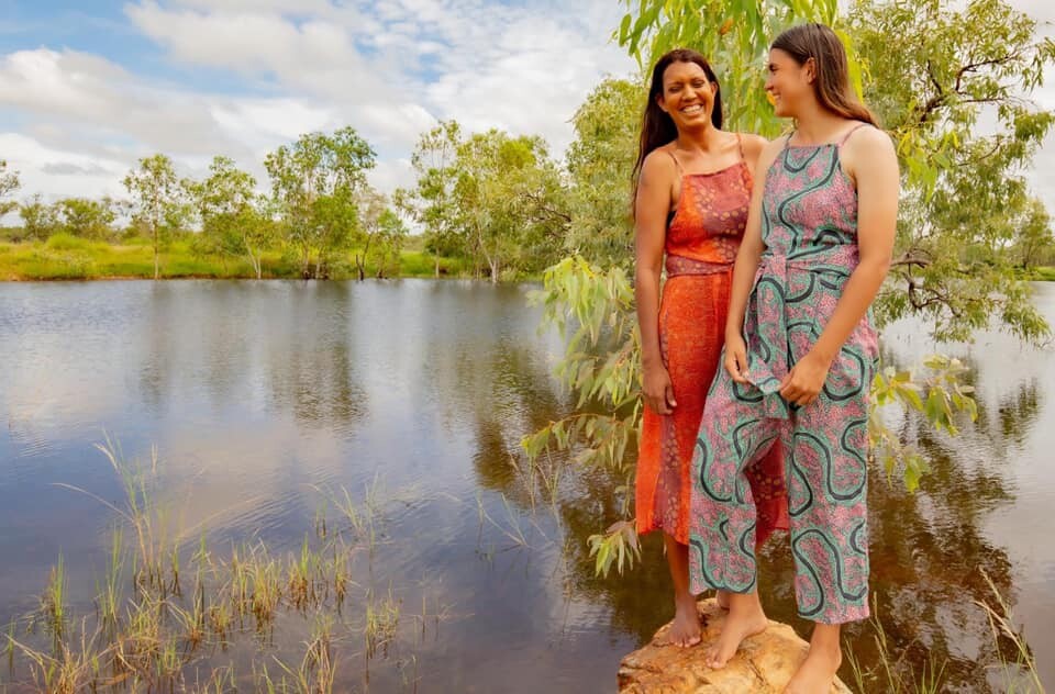 Two girls stand by a river, smiling as they model the Jaru Girl fashion line