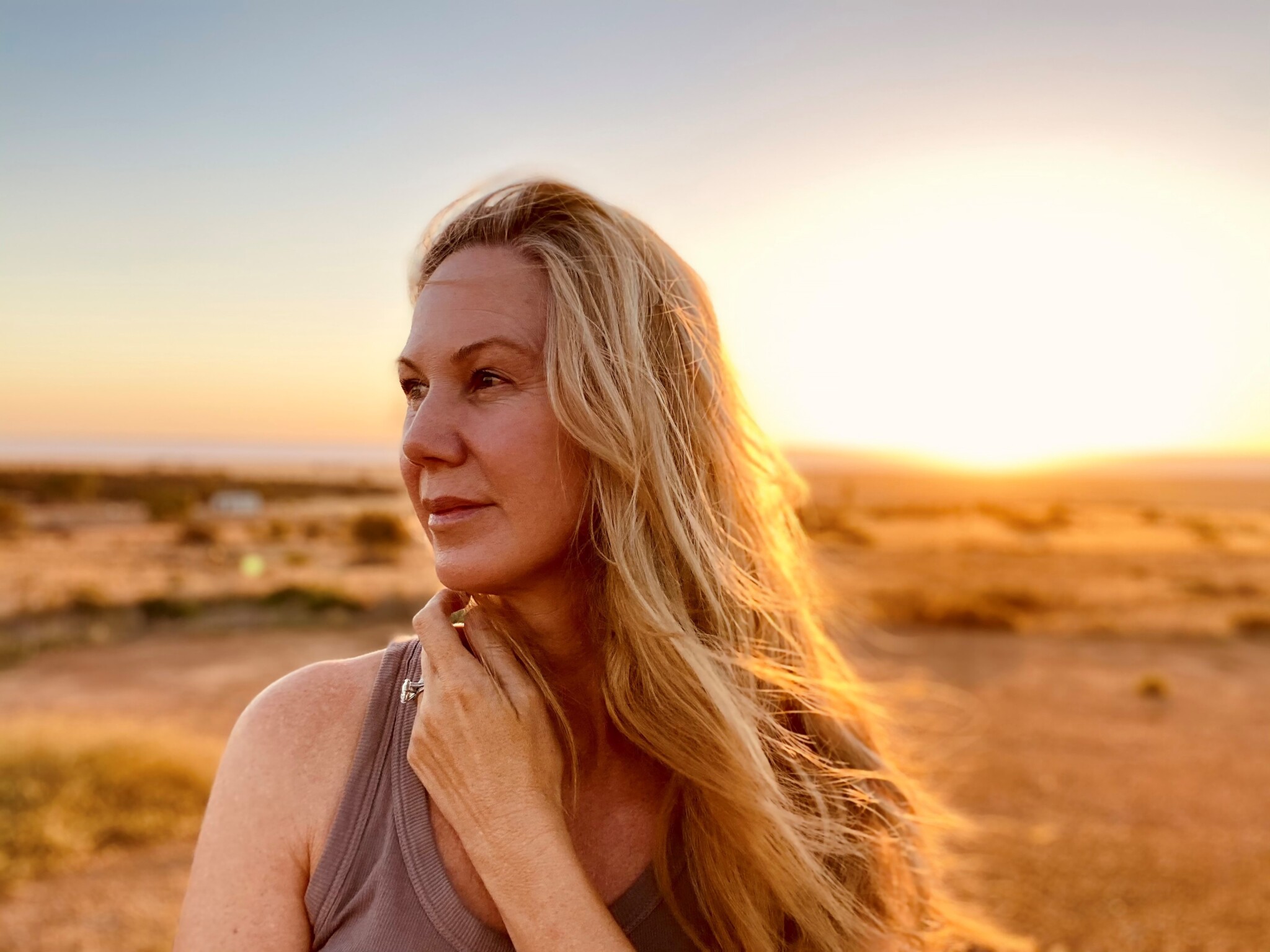 Lynda Howitt standing in a field. Lynda has long blonde hair and is wearing a grey singlet. She is looking into the distance and holding her arm is crossed over so her hand sits under her chin. The image is backlit with golden light.