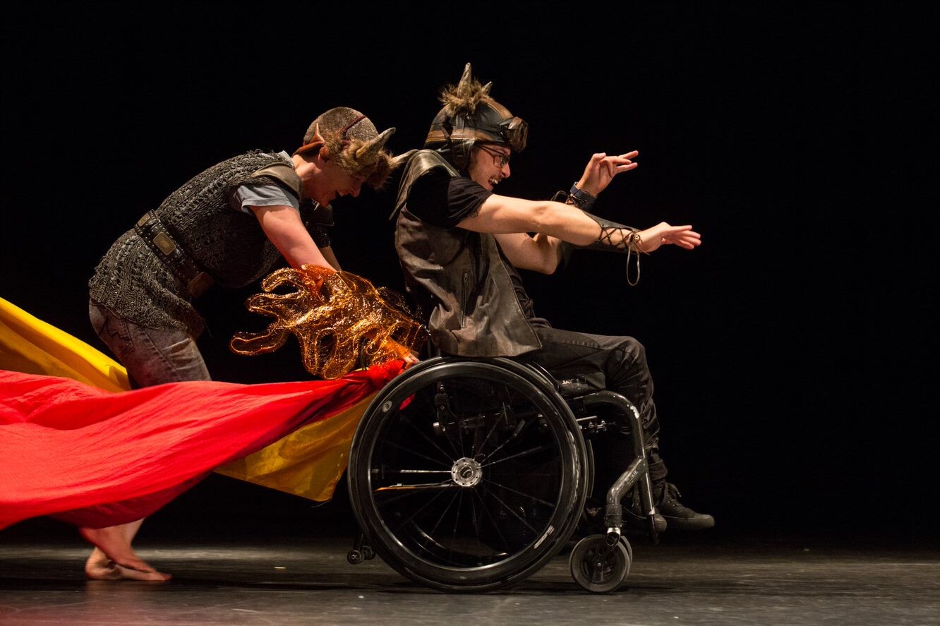 Two people on stage, one is in a wheelchair wearingarm braces, being pushed by another wearing horns on their head.