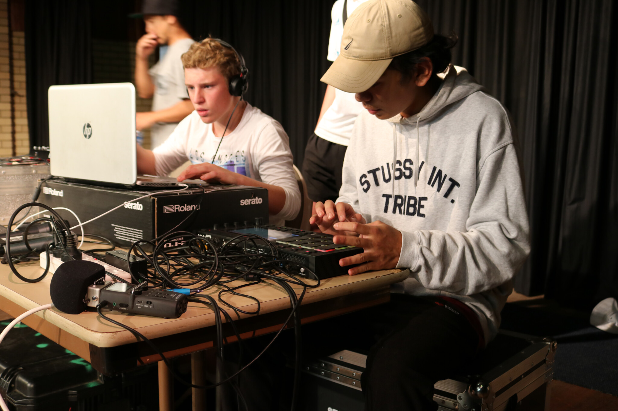 Two individuals working with audio equipment; one uses laptop with headphones, other adjusts device with buttons; cables and electronics on table.