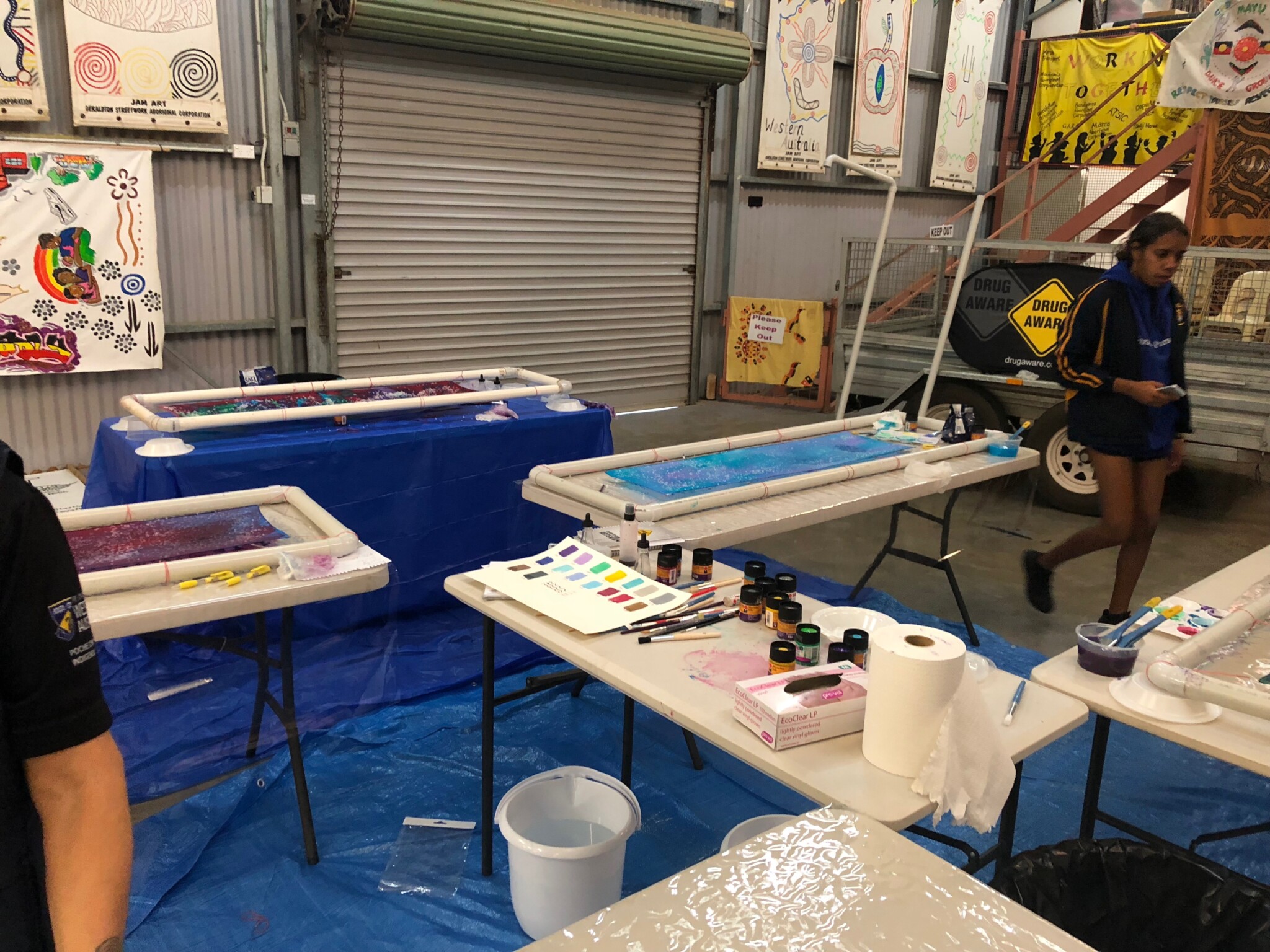 Indoor art workshop with tables set up for painting; supplies include paint jars, brushes, trays with liquid; artworks on walls, blue floor sheeting.