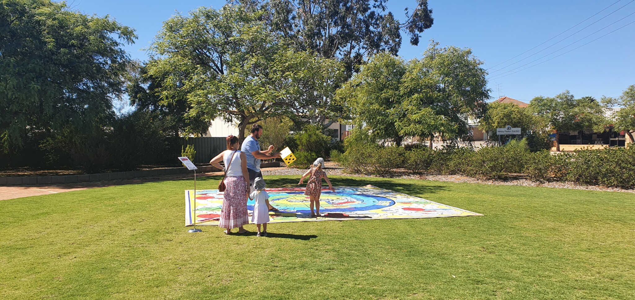 Group of adults and children standing on large colorful game mat in grassy park; one person holds large yellow die; trees and buildings behind.