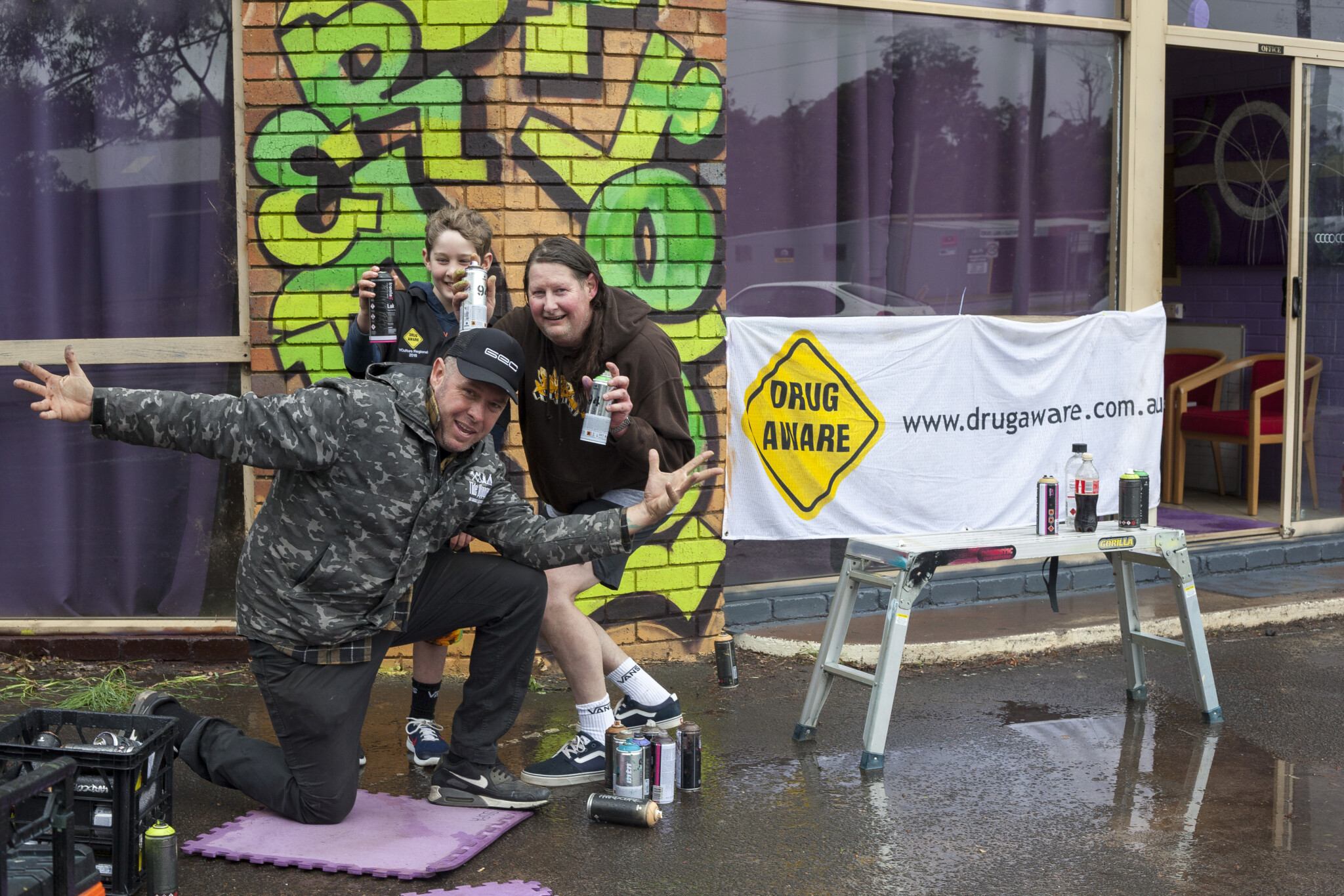 Three people pose in front of graffiti-covered brick wall; holding spray paint cans near "DRUG AWARE" banner and table.