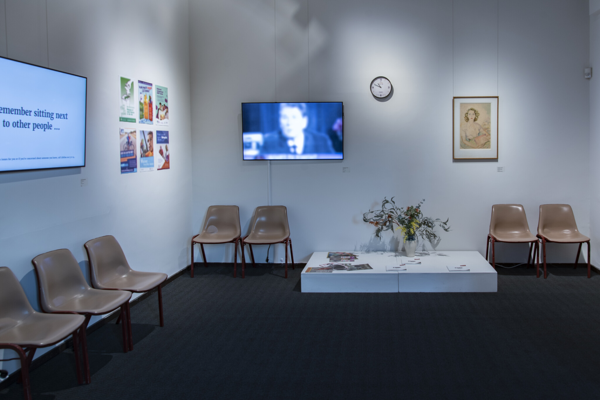 Gallery set up to look like a hospital waiting room. There are chairs around the walls in sets of two and three, and two TV screens on the wall. One TV screen reads, 
