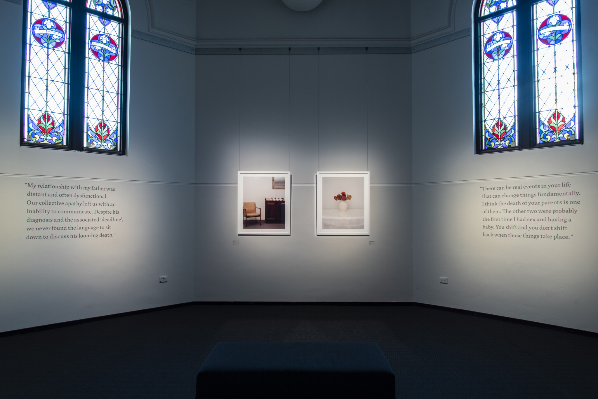 Two photos hung on wall. One is of a chair, one is of a flower vase. Descriptive text is written on the walls to the right and left. Two large stained-glass windows sit above the text.