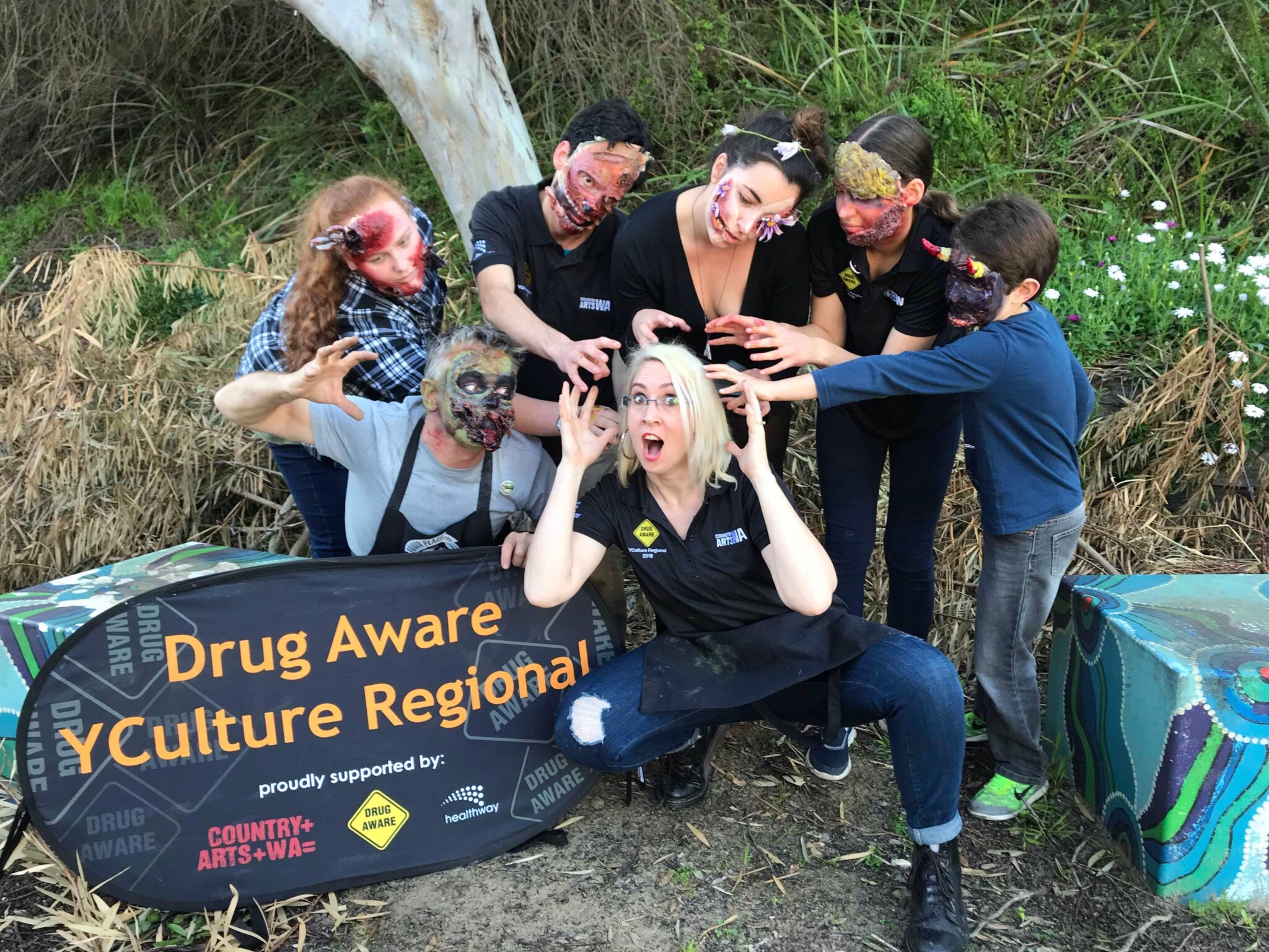 Group of people posing outdoors with partially obscured faces, holding "Drug Aware YCulture Regional" banner