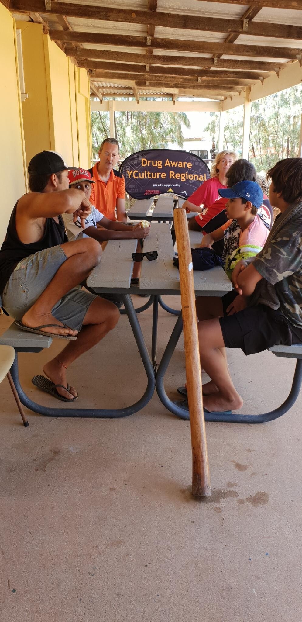 Group of people seated around rectangular table under covered outdoor area; "Drug Aware YCulture Regional" sign visible in background.