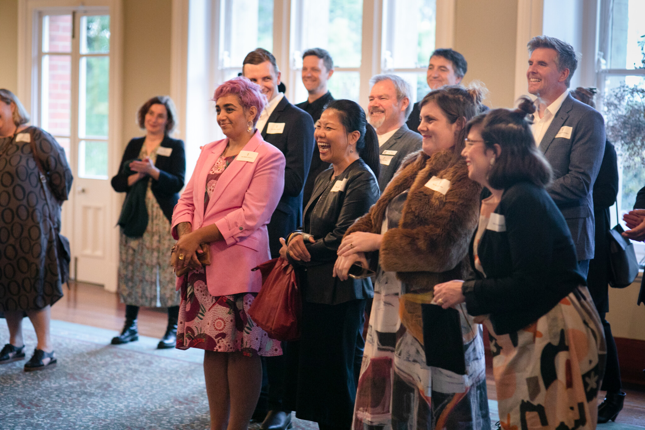 Group of people indoors at formal event; dressed in suits, dresses, and coats; some wear name tags; large windows in background.