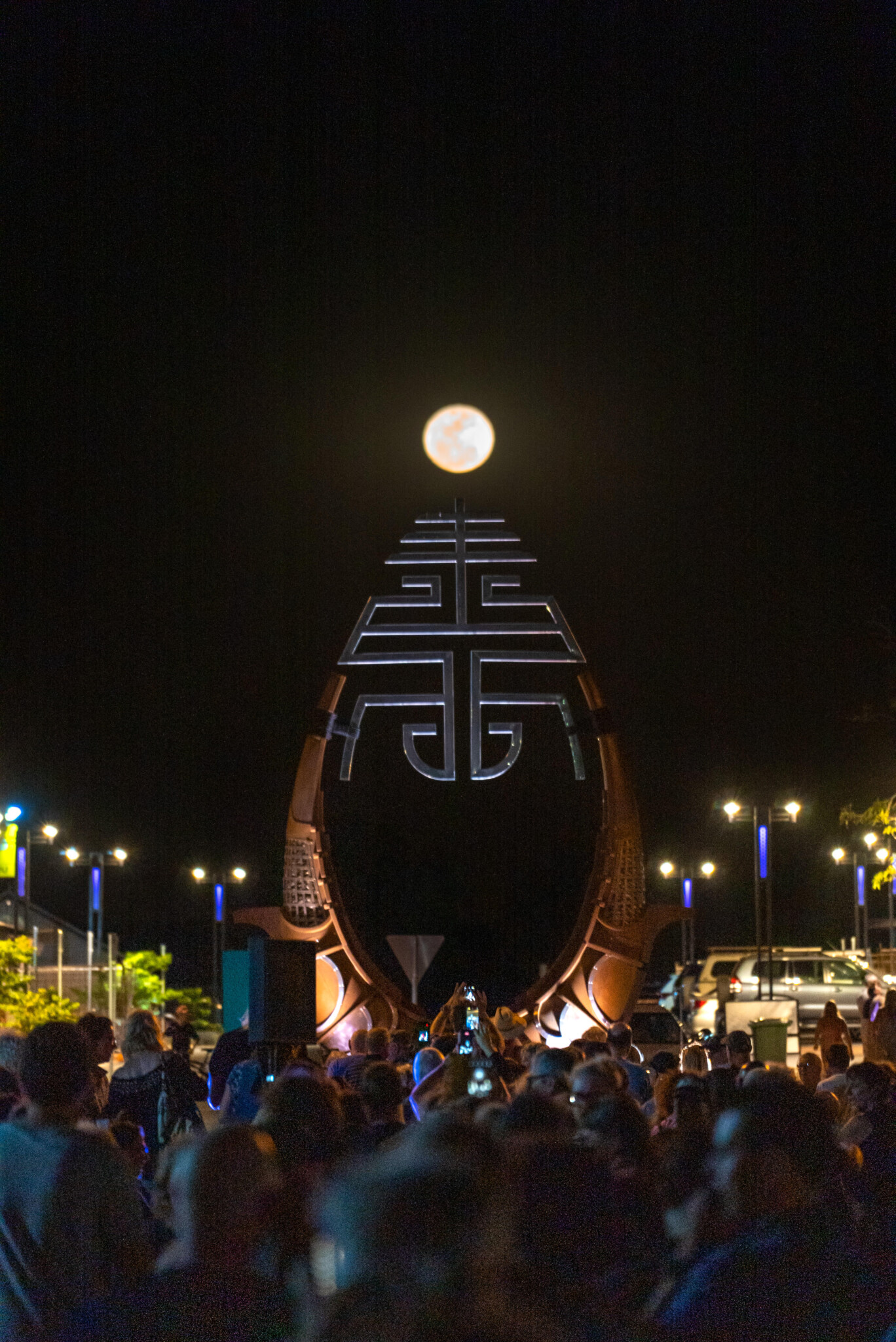 Night crowd gathers before tall lit sculpture aligned with full moon; geometric patterns glow; streetlights in background.