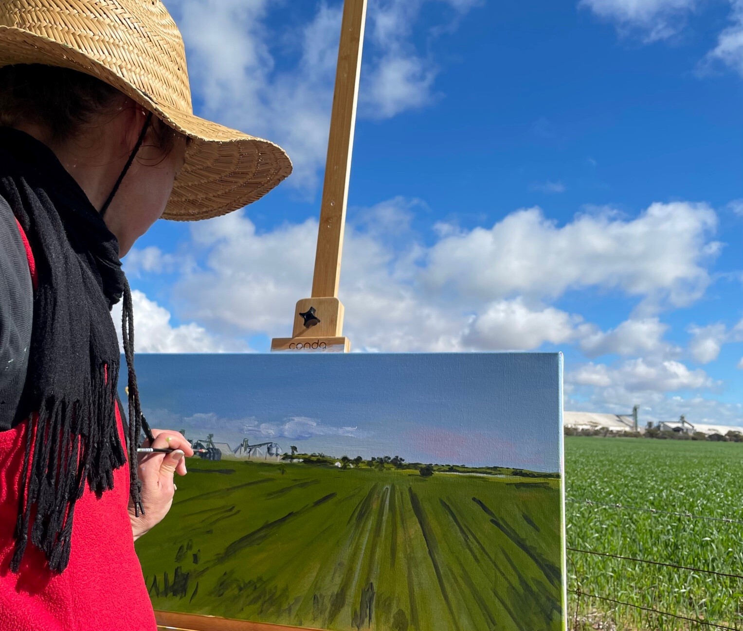 Artist Siobhan Kelley working on a plein air painting of a wheat field and blue sky. Siobhan is in the foreground and is wearing a black and red jumper and straw hat while she paints a canvas on an easel.