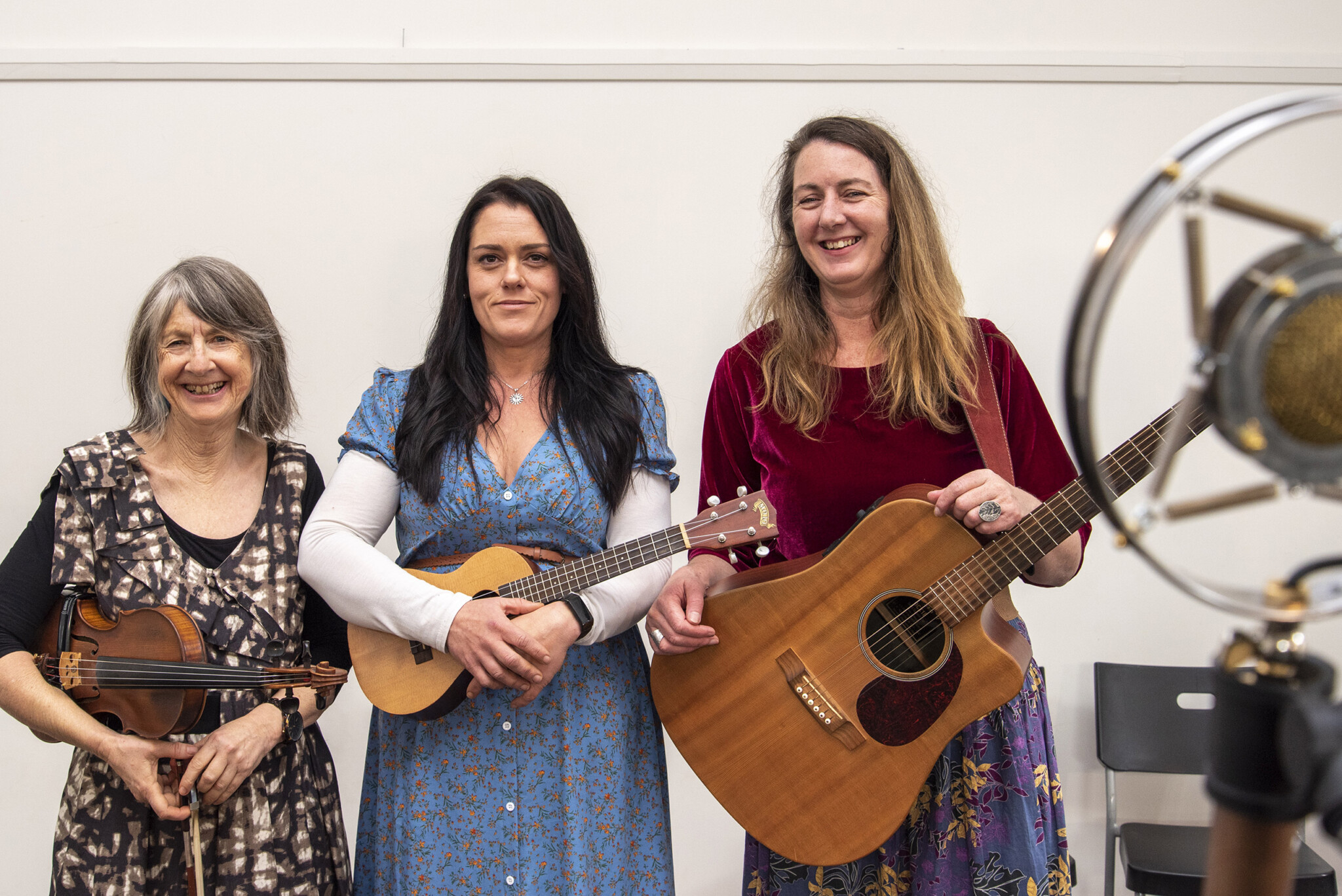 Three women standing in height order. The first, an older lady with grey hair in a dark patterned dress holds a violin. The middle woman with dark brown hair in a blue dress holds a ukelele and the third woman with blonde hair holds a guitar.