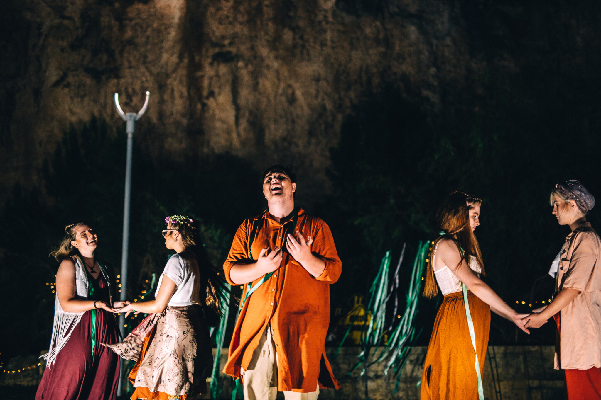 Pairs of people in colorful attire hold hands at night outdoor event; rocky cliff and light post in background.