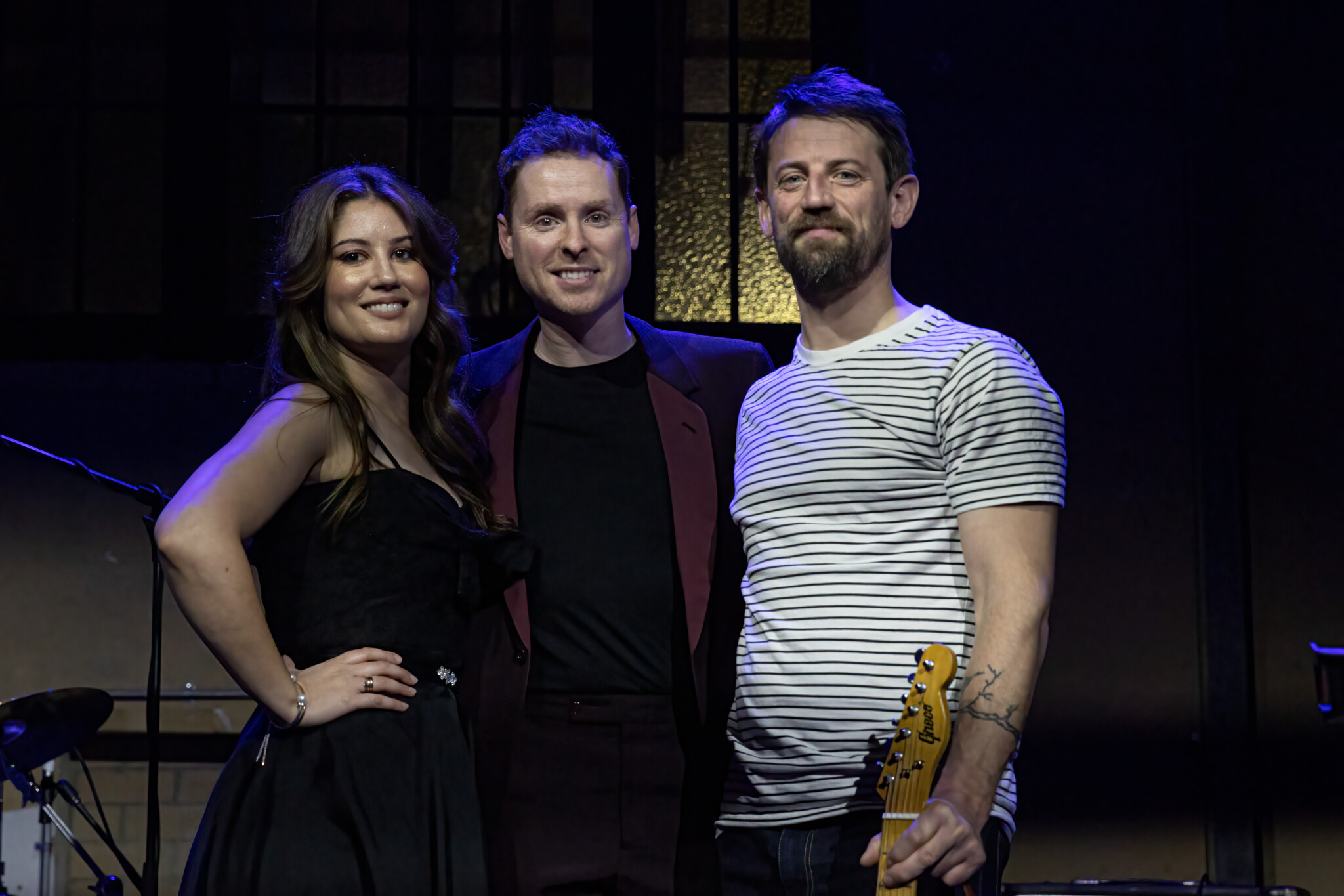Three people stand on stage, one holds a guitar.