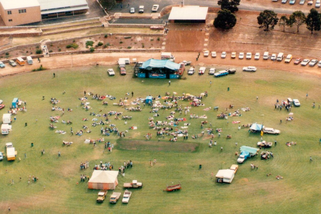 Vintage aerial photo. Crowds of people are scattered over a large football oval. There is a blue marquee at one end.