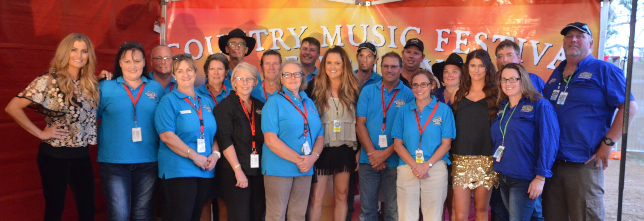Approximately 21 people stand grouped together in front of the Country Music Festival Banner. Most of them wear blue polo shirts and have red lanyards around their neck.
