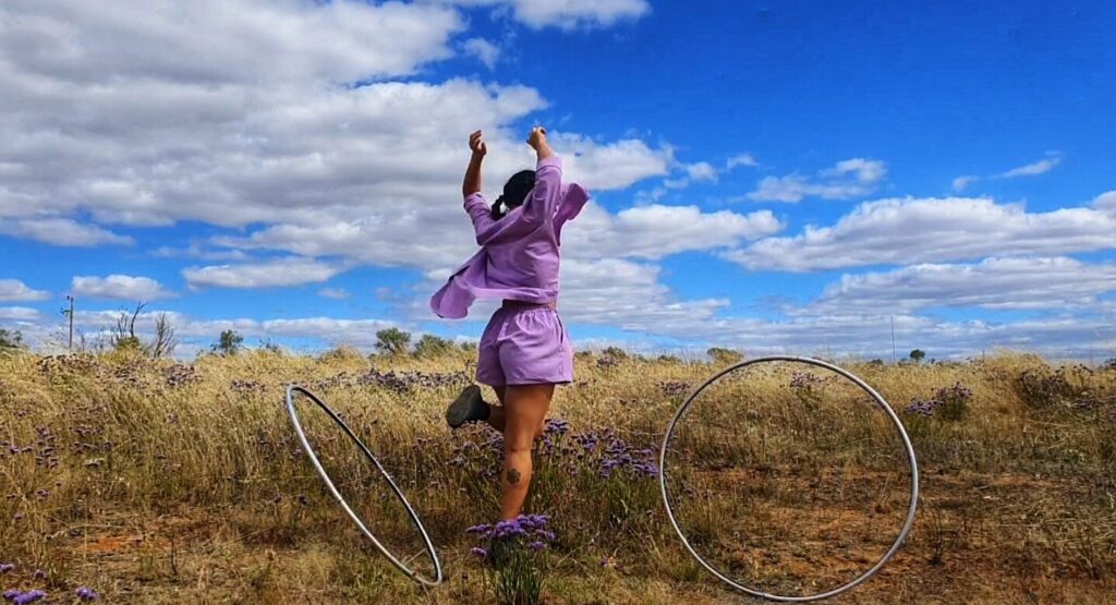 A woman stands in a field of grass and purple flowers. She is wearing lavender purple shorts and matching button up top. She is facing away from the camera and has her arms are in the air and her left knee is bent. There is a small tattoo on her right calf. There are two hula hoops either side of her.
