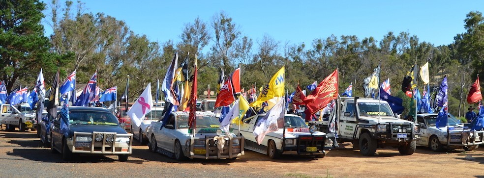Many utes and trucks parked on a dirt road. They are all adorned with flags. Many are the Australian flag, others include flags for breast cancer awareness, and others are unidentifiable