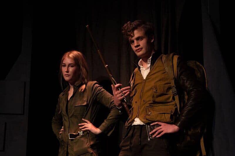 Two people stand side by side with faces obscured; one in green outfit with belt, other in vest holding rifle.