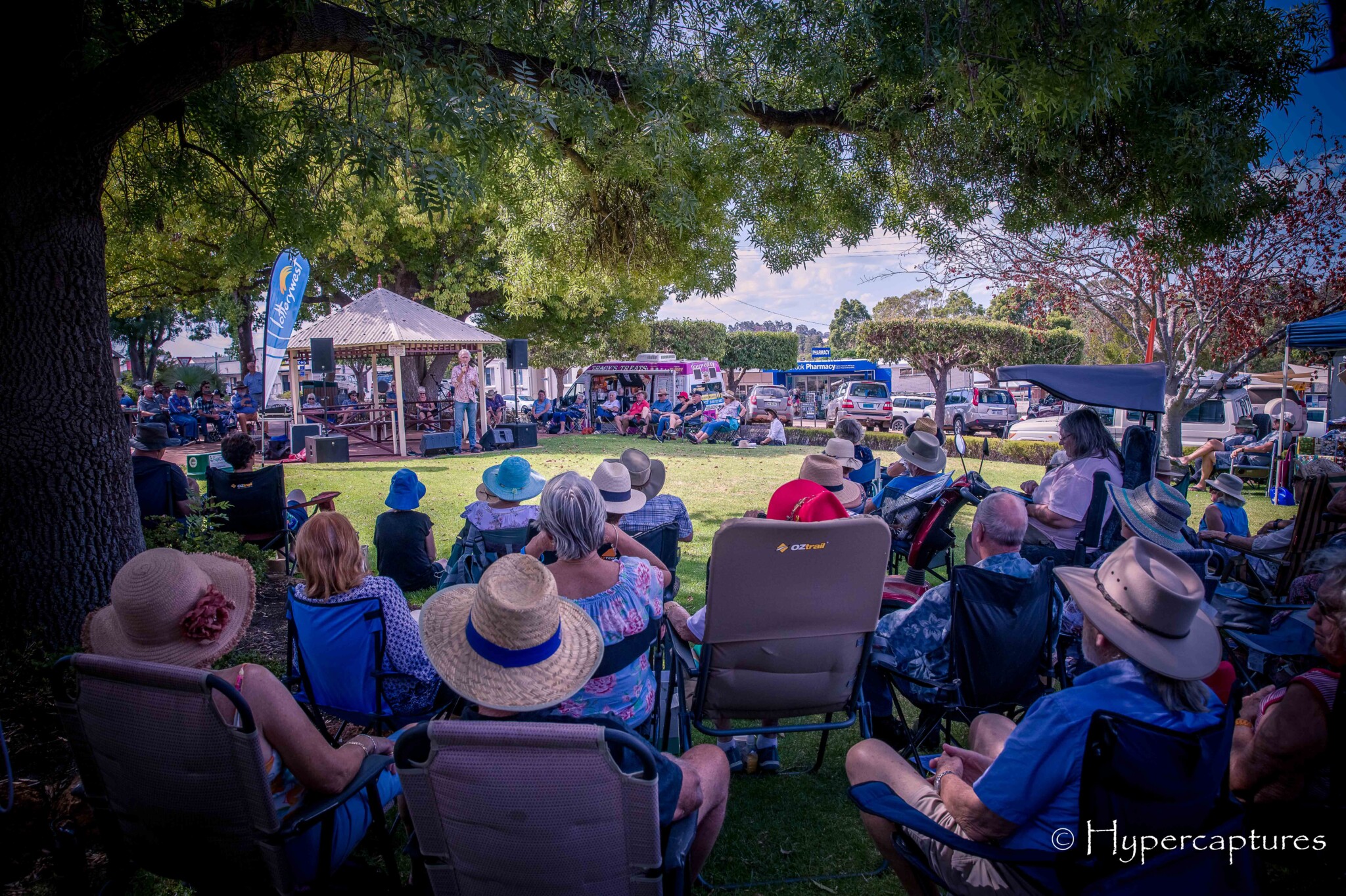 Large group of people sitting in chairs under the tree of a park. There is a woman speaking into a microphone under a marquee in front of them.