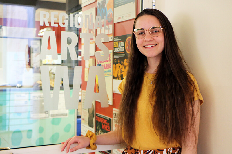 Person with long brown hair standing next to a door that reads "REGIONAL ARTS WA"