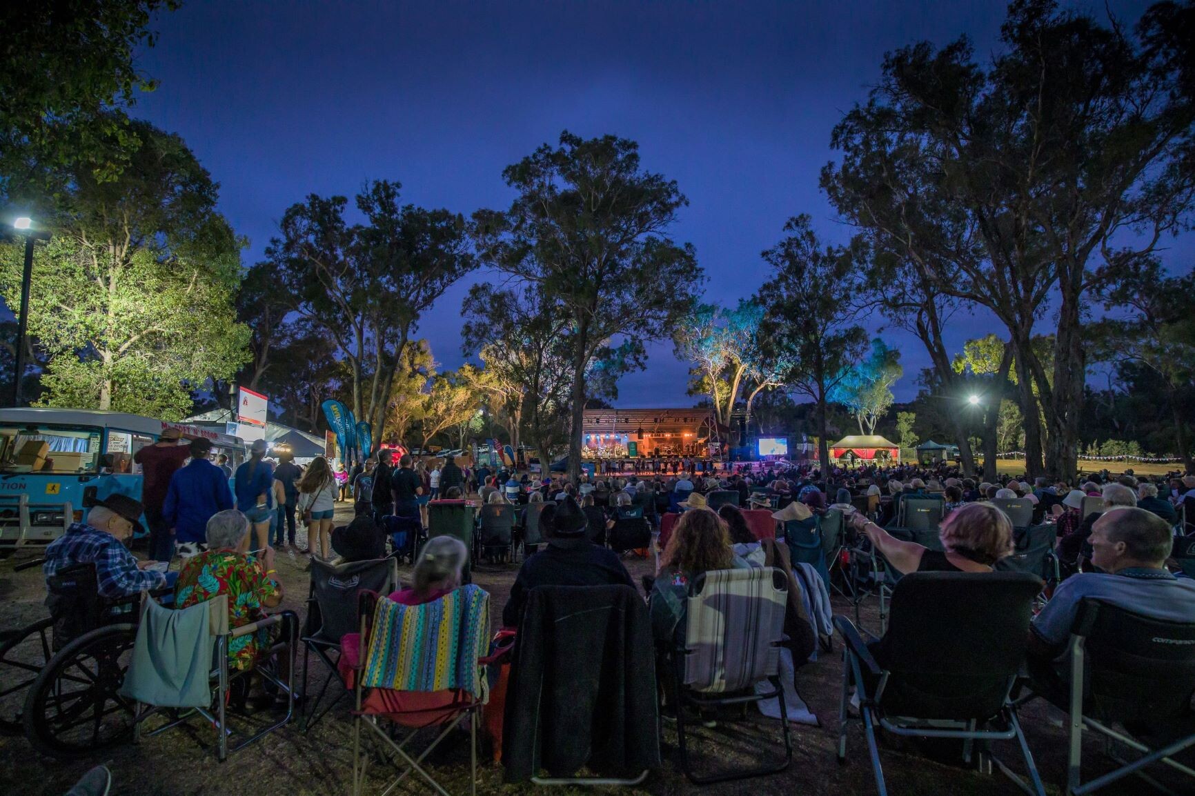 Large outdoor evening event with audience in chairs and wheelchairs facing lit stage; tents and tall trees in background.