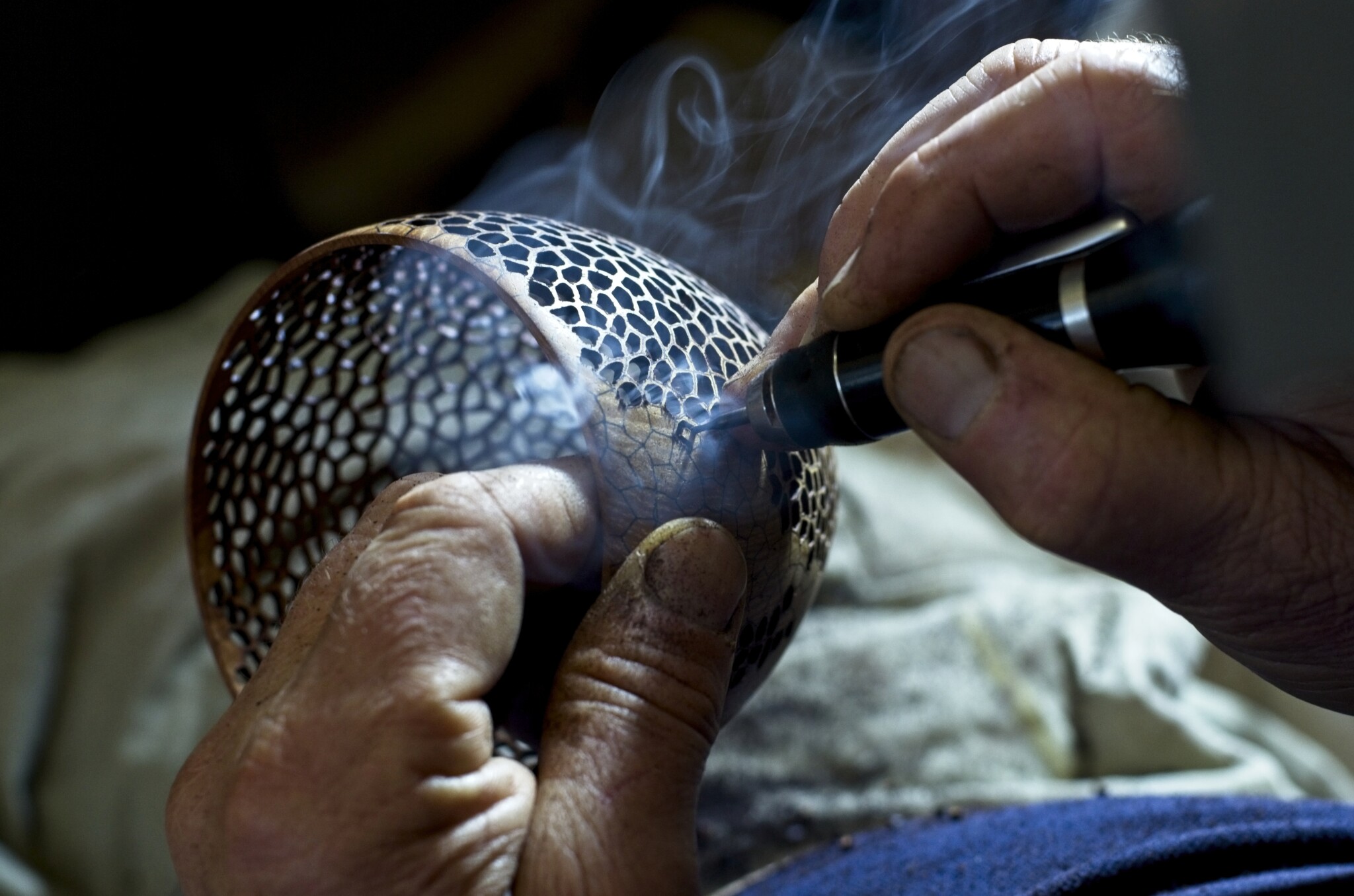 Close-up of Neil creating delicate perforations in a wooden vase. The tool is smoking.