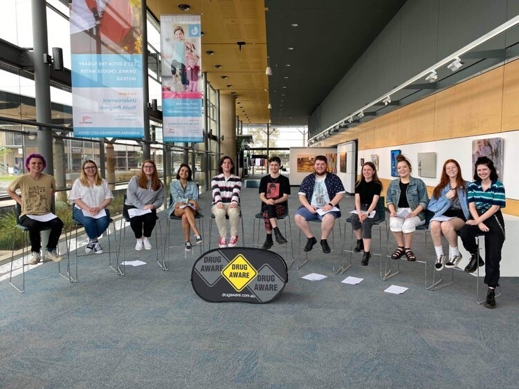 Eleven people sit on chairs, in a row in front of the yellow "Drug Aware" Banner.