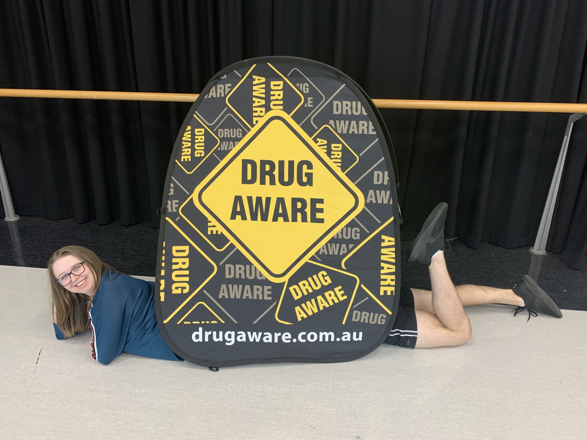 Two young people lay inside a large banner reading Drug Aware. The girl at the front has her head sticking out and smiles at the camera, there is a boy at the back who sticks his legs out the back, with one raised. This gives the illusion of a very long person with mismatched head/legs